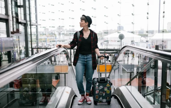 A person with a suitcase stands on an escalator in an airport, wearing casual clothes, amidst modern architecture and busy travelers below.