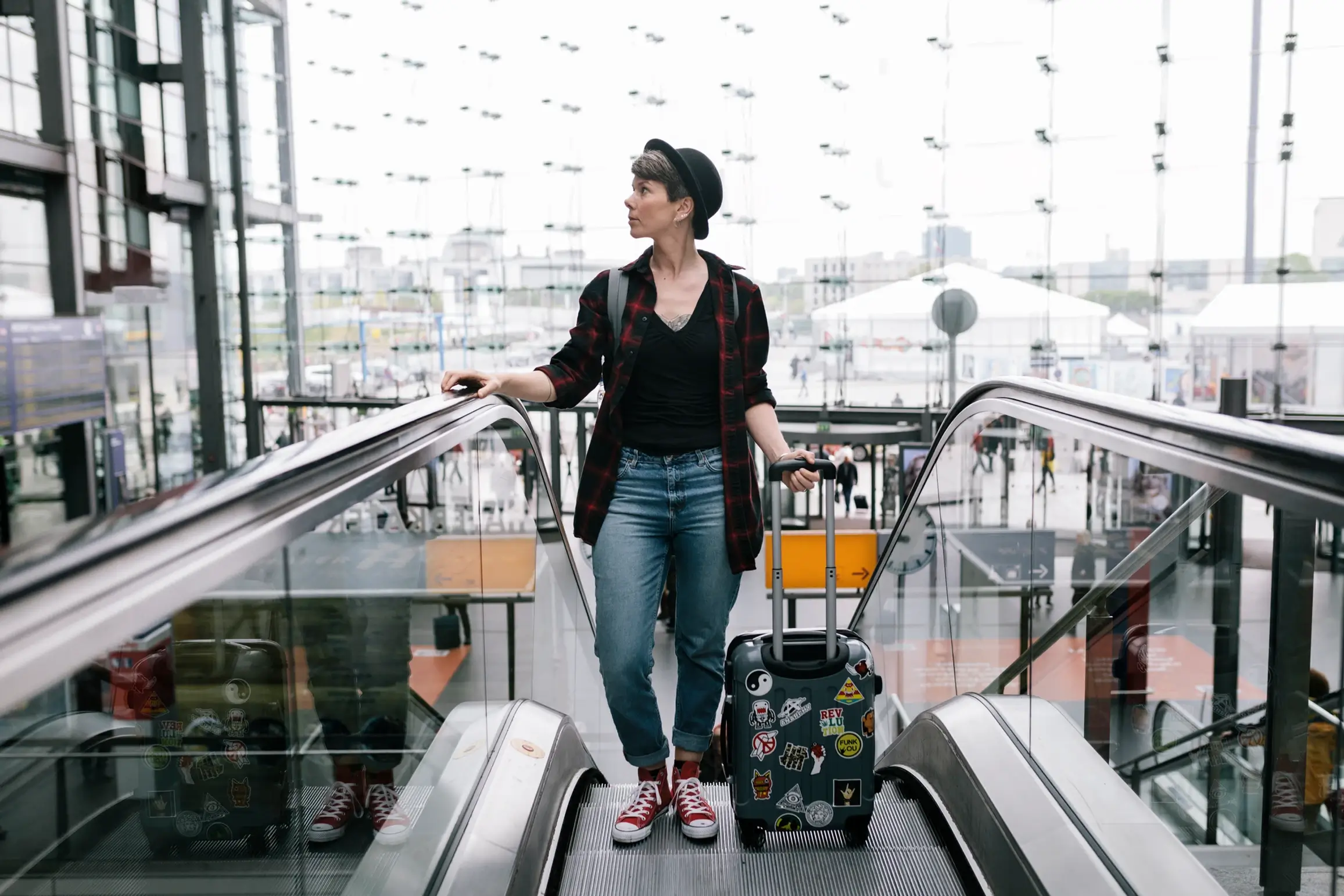 A person with a suitcase stands on an escalator in an airport, wearing casual clothes, amidst modern architecture and busy travelers below.