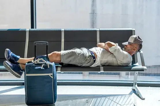 A person is sleeping on an airport bench, wearing casual clothes and a cap. A suitcase is placed nearby under sunlight.