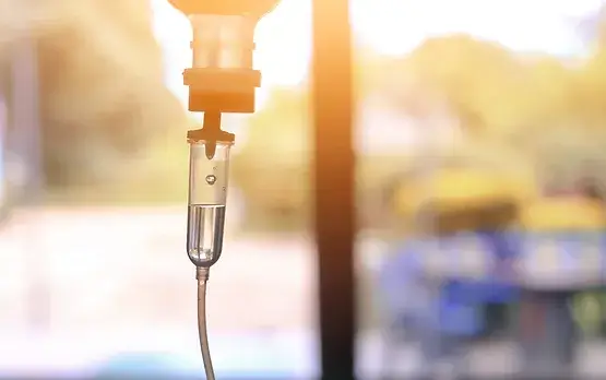 Close-up of an IV drip with liquid, set against a softly blurred, sunlit background, suggesting a medical environment or hospital setting.