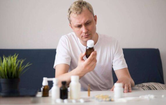 A person in a white shirt examines a medicine bottle, surrounded by several other bottles and pills on a table indoors.