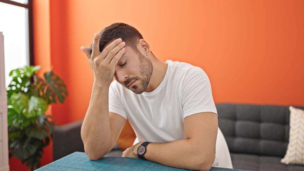 A person in a white shirt rests their head on a hand, sitting at a table, with an orange backdrop and plants nearby.
