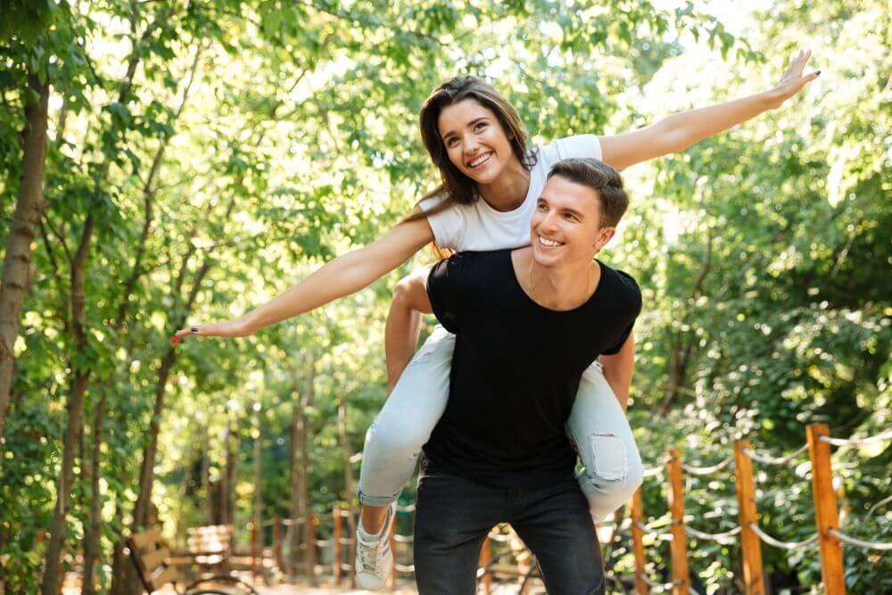 Two people enjoying a playful piggyback ride on a sunny day in a lush, tree-lined park with benches.